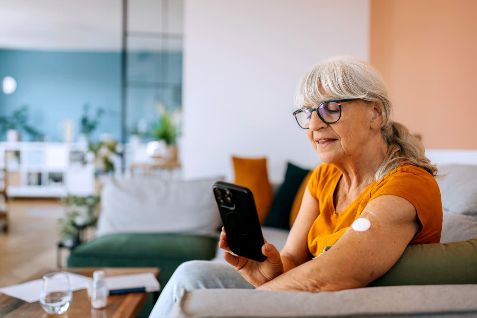 An older woman relaxing on her couch, checking her glucose readings on her phone — a CGM sensor on her arm